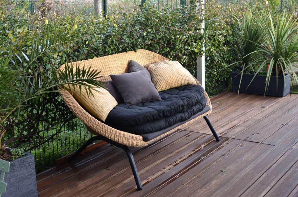 Comfortable stylish wicker sofa with soft seat and pillows placed in patio with lush green tropical vegetation in daytime